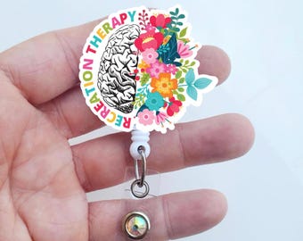 Op de afbeelding: Een witte plastic badgehouder met een transparante intrekbare rol. De badgehouder heeft een kleurrijk bloemenontwerp met een hersenafbeelding en de tekst "Recreatietherapie".