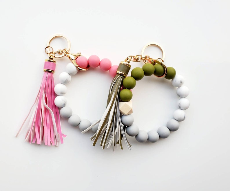 May include: A set of two beaded keychains with tassels. One keychain has pink and white beads with a pink tassel. The other keychain has green and white beads with a green and white tassel.