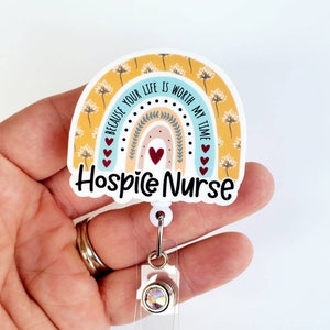May include: A yellow, blue, and pink rainbow with a white background and the text "Because your life is worth my time" and "Hospice Nurse". The rainbow has a white border and is attached to a clear plastic retractable badge holder with a silver clip and a clear rhinestone.