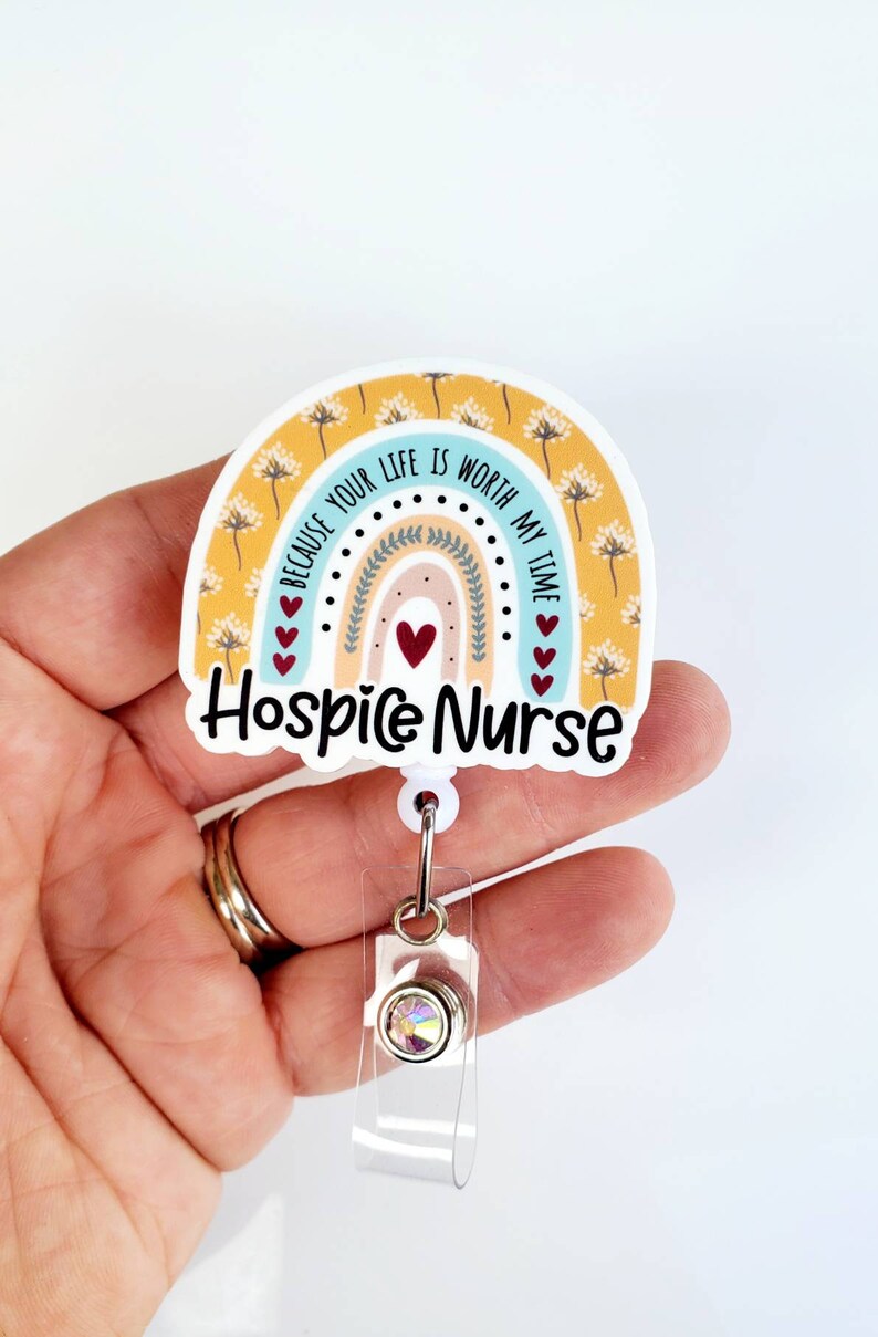 May include: A yellow, blue, and pink rainbow with a white background and the text "Because your life is worth my time" and "Hospice Nurse". The rainbow has a white border and is attached to a clear plastic retractable badge holder with a silver clip and a clear rhinestone.