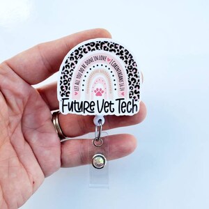 May include: A white and pink rainbow with a paw print and the text "Let all you do be done in love 1 Corinthians 16:14" and "Future Vet Tech" on a leopard print background. The rainbow is attached to a clear retractable badge holder with a silver chain and a clear rhinestone.