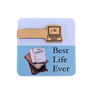 May include: Wooden bookmark with a laptop design and the initials "J.W." engraved on it. The bookmark is placed on a blue background with a stack of books and the text "Best Life Ever".