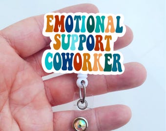May include: A white badge holder with a colorful sticker that reads "Emotional Support Coworker". The sticker has a retro font style and is in shades of blue, green, orange, and red. The badge holder has a clear plastic cover and a silver clip.