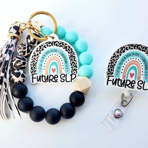 May include: A keychain and badge reel set with a teal and black leopard print rainbow design. The keychain has a black tassel and black and white beads. The badge reel has a clear plastic holder and a silver clip. Both items feature the text "Let all you do be done in love I Corinthians 16:14" and "Future SLP".
