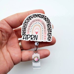 May include: A white and pink rainbow with a leopard print border. The rainbow has a pink heart in the centre and the text "Let all you do be done in love 1 Corinthians 16:14" around the outside. The text "APRN" is written below the rainbow. The rainbow is attached to a clear retractable badge holder with a pink rhinestone.
