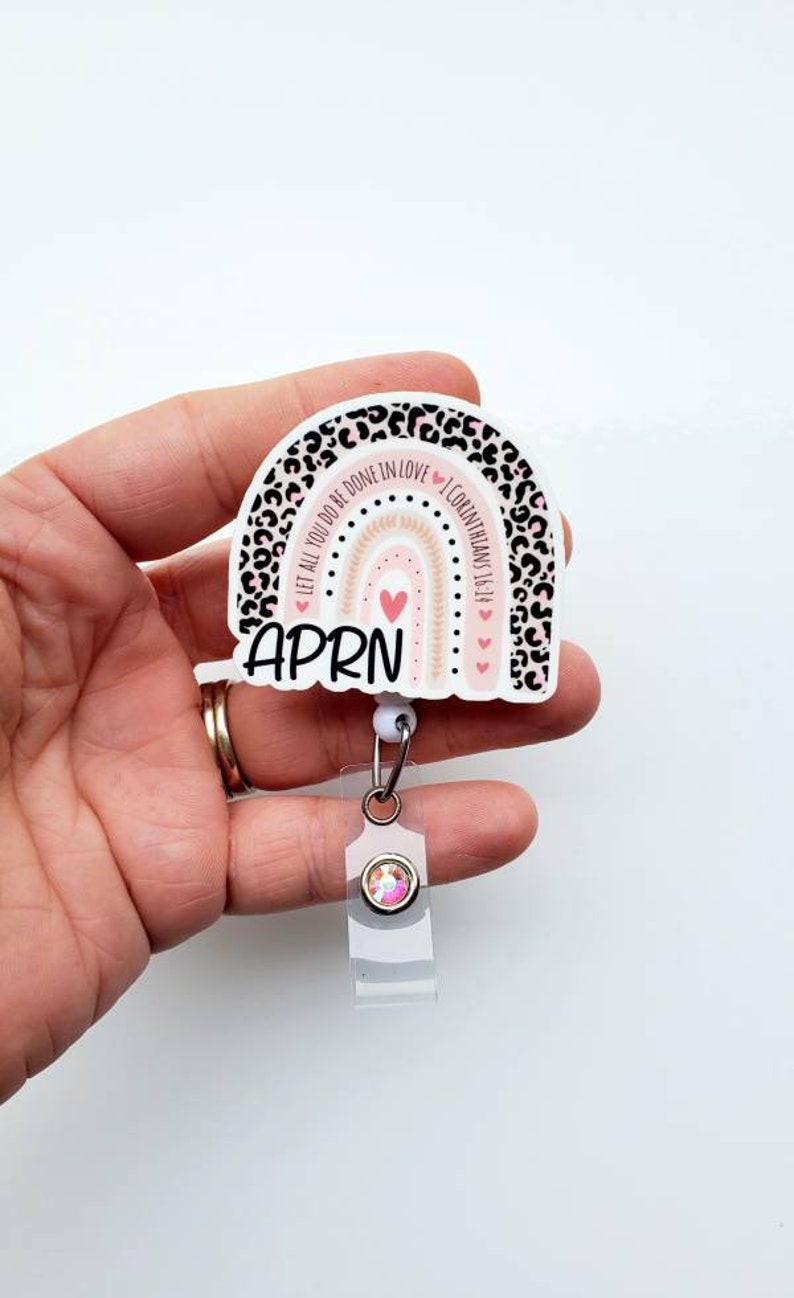 May include: A white and pink rainbow with a leopard print border. The rainbow has a pink heart in the centre and the text "Let all you do be done in love 1 Corinthians 16:14" around the outside. The text "APRN" is written below the rainbow. The rainbow is attached to a clear retractable badge holder with a pink rhinestone.