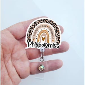 May include: A white and brown leopard print rainbow with the text "Let all you do be done in love 1 Corinthians 16:14" and the word "Phlebotomist" below. The rainbow is attached to a clear retractable badge holder with a sparkly rhinestone.