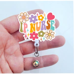 May include: A white plastic badge holder with a colorful design featuring the text "IP NURSE" surrounded by flowers and hearts. The badge holder has a clear plastic retractable cord and a crystal-like button.