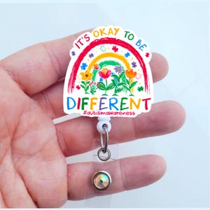 May include: A white retractable badge holder with a colorful rainbow and flower design. The design says "It's okay to be different" and "#autism awareness".