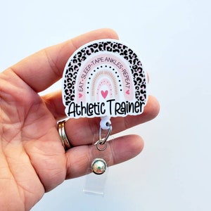 May include: A white and pink retractable badge holder with a leopard print border. The badge holder features a rainbow with the text "Eat Sleep Tape Ankles Repeat" and the words "Athletic Trainer".