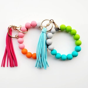May include: Two colorful silicone beaded keychains with tassels. One keychain is pink, orange, and gray, and the other is green, blue, and gray.