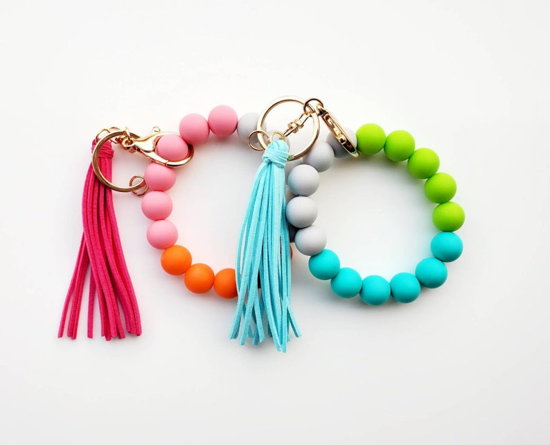 May include: Two colorful silicone beaded keychains with tassels. One keychain is pink, orange, and gray, and the other is green, blue, and gray.