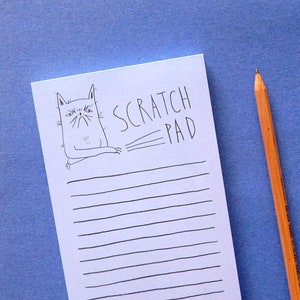 May include: A white scratch pad featuring a black cat illustration and the words "SCRATCH PAD". A yellow and orange pencil with a pink eraser is placed on a blue surface.