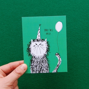 May include: A green card with a black and white illustration of a cat wearing a party hat and holding a white balloon. The text "YOU'RE OLD." is printed on the card.