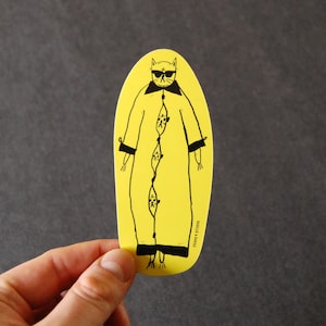 May include: Yellow sticker with a black and white illustration of a cat wearing sunglasses and a long robe. The cat has a long tail and is standing with its arms at its sides. The text "Regina & Rose" is printed on the bottom right corner of the sticker.