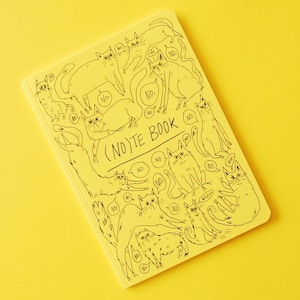 May include: A yellow notebook with a black illustration of cats and the text "(NO)TE BOOK". The cats are in various poses, with speech bubbles saying "NO". The notebook is on a yellow background.
