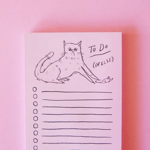 May include: A pink to-do list notepad with a black and white illustration of a cat with a grumpy expression chasing a mouse. The text on the notepad says "To Do (or else)".