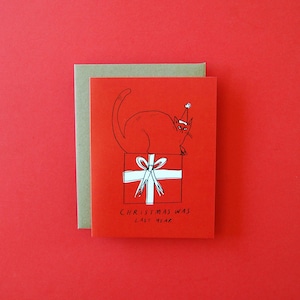 May include: A red and white Christmas card with a black line drawing of a cat wearing a Santa hat sitting on top of a wrapped present. The text on the card reads "Christmas was last year."