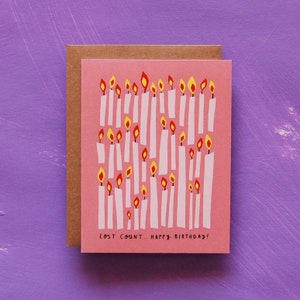 May include: Pink birthday card with a white envelope. The card features a pattern of white candles with yellow and red flames. The text on the card reads "Lost count... Happy Birthday!"