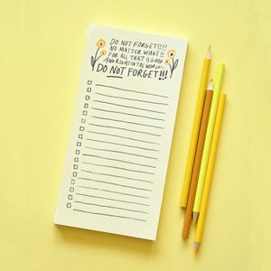 May include: A rectangular notepad with a cream-coloured paper surface featuring black hand-drawn lines, checkboxes, and the text DO NOT FORGET NO MATTER WHAT FOR ALL THAT IS GOOD AND RIGHT IN THE WORLD DO NOT FORGET, accompanied by three yellow pencils.