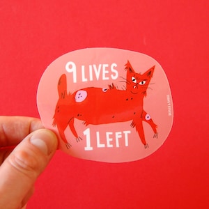 May include: A red sticker with a cartoon cat illustration. The cat has a grumpy expression and is covered in spots. The text on the sticker reads "9 LIVES 1 LEFT". The sticker is held by a hand against a red background.