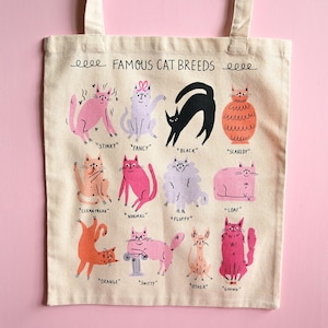 May include: A beige canvas tote bag featuring a graphic print titled FAMOUS CAT BREEDS. The design displays twelve illustrated cats in various colors and poses, each labeled with descriptive terms like Stinky, Fancy, Black, Scaredy, Clean Freak, Normal, Fluffy, Loaf, Orange, Snitty, Other, and Giving.