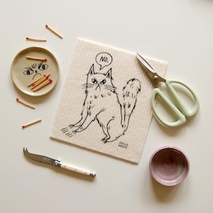 May include: A whimsical still life featuring a beige cat-themed cleaning cloth with the word "NO" in a speech bubble. Accompanying items include a small ceramic dish with a grumpy face, matches, scissors, a cheese knife, and a small pink bowl.