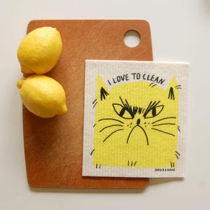 May include: A white dishcloth with a yellow cat face and the text "I love to clean." The cat has black eyes and a black nose. The dishcloth is on a brown wooden cutting board with two yellow lemons.
