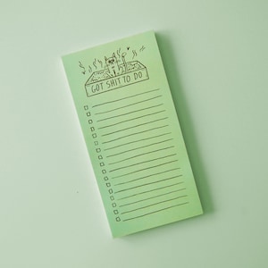 May include: A rectangular, light green notepad with a black and white cartoon cat illustration above the text "GOT SHIT TO DO." The notepad has horizontal lines for writing and check boxes. The cat is in a litter box.