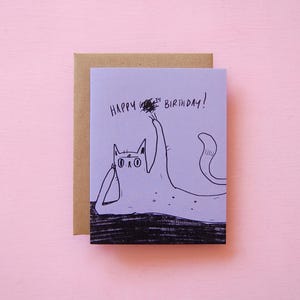 May include: A lilac-colored birthday card with a hand-drawn illustration of a cat. The cat is drawn in black ink and is holding a small object. The words "HAPPY BIRTHDAY!" are written at the top of the card. The card is paired with a brown envelope.