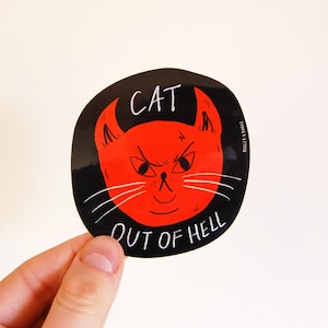 May include: A black sticker featuring a red cat face with devil horns and a grumpy expression. White text reads "CAT OUT OF HELL". The sticker is held by a hand. The design is a humorous take on a cat.