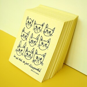 May include: A stack of pale yellow rectangular cleaning cloths featuring a black line drawing of nine cat faces with grumpy expressions. The text reads, "i'm not mad, i'm just disappointed!" followed by the brand name "BADGER & BURKE" at the bottom.