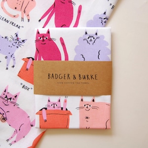 May include: A white tea towel with a colorful cat design. The towel features a variety of cats in different poses, including a cat in a box, a cat with a crown, and a cat with a fish. The text "LEAN FREAK", "BOX", and "BADGER & BURKE 100% COTTON TEA TOWEL" is printed on the towel.