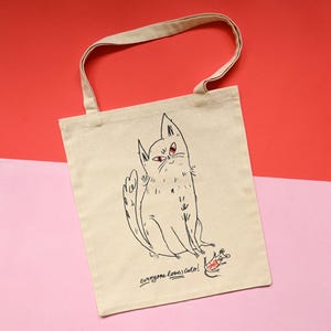 May include: A beige canvas tote bag featuring a black line drawing of a cat with red eyes and a small mouse with a red bow. The text "everyone loves cats!" is printed in black script below the cat illustration. The bag has two shoulder straps.