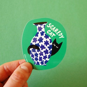 May include: A green sticker with the words "Scaredy Cat" and a black cat peeking out from behind a blue and white floral vase. The sticker is held by a hand against a green background.