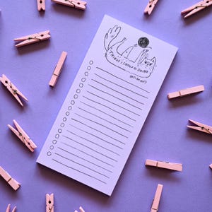 May include: A rectangular light purple notepad featuring a black ink illustration of a cat playing with a ball of yarn. The text reads "THINGS I SHOULD BE DOING (BUT I AM NOT)" above a vertical list of blank lines with circular checkboxes.