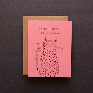 May include: A pink Valentine's Day card with the text "You'll do... Happy Valentine's Day." The card features a black illustration of a spotted cat holding a heart. The card is inside a brown envelope.
