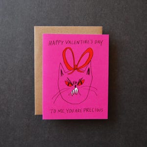 May include: A pink Valentine's Day card with a drawing of an angry cat wearing a red bow. The text "HAPPY VALENTINE'S DAY" and "TO ME, YOU ARE PRECIOUS" are printed on the card. The card is paired with a brown envelope.