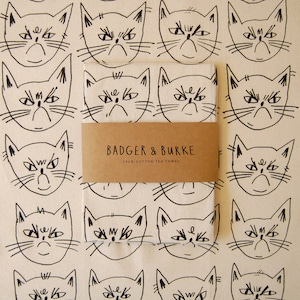 May include: A white tea towel with a repeating pattern of black line drawings of grumpy cats. The text "BADGER & BURKE 100% COTTON TEA TOWEL" is printed in brown on a brown label.