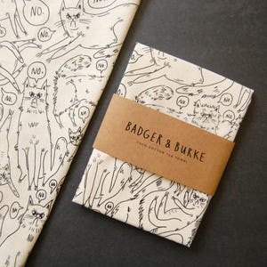 May include: A white tea towel with a black and white illustration of cats saying "no". The tea towel is packaged with a brown paper label that reads "Badger & Burke 100% Cotton Tea Towel".