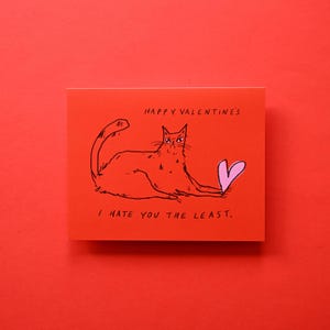 May include: A rectangular greeting card with a bright red background featuring a black ink illustration of a grumpy cat holding a pink heart. The text reads HAPPY VALENTINE'S at the top and I HATE YOU THE LEAST. at the bottom in black ink.