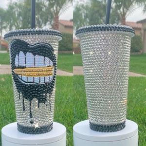 May include: Two clear plastic tumblers with black lids and straws. One tumbler is covered in rhinestones and features a black and gold mouth with teeth design. The other tumbler is covered in rhinestones.