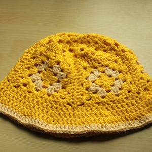 May include: A yellow crocheted bucket hat with a beige trim. The hat has a textured pattern with squares and circles.