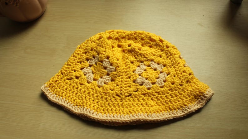 May include: A yellow crocheted bucket hat with a beige trim. The hat has a textured pattern with squares and circles.