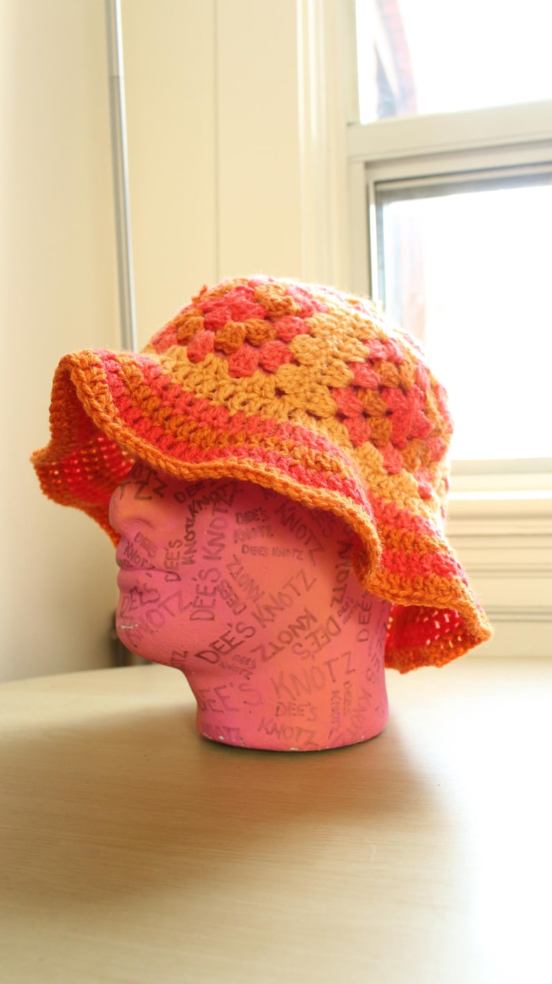 May include: A crocheted sun hat with a wide brim in orange and pink. The hat is on a pink mannequin head.
