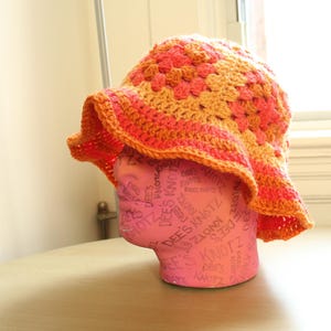 May include: A crocheted sun hat with a wide brim in shades of orange and pink. The hat is made with a granny square pattern.