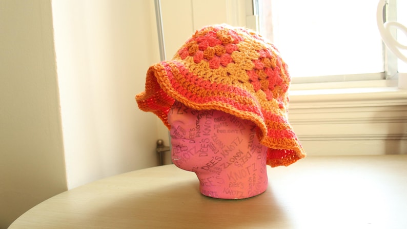 May include: A crocheted sun hat with a wide brim in shades of orange and pink. The hat is made with a granny square pattern.