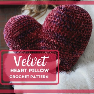 May include: A close-up of a red velvet heart-shaped pillow. The pillow is being held by a person wearing a white sweater. The text "Velvet Heart Pillow Crochet Pattern" is written in white on a red background.