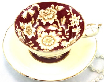 May include: A red teacup and saucer with a white floral pattern and gold trim. The cup has a scalloped rim and a handle. The saucer is plain white with a gold rim.