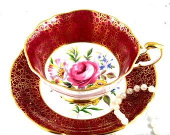 May include: A vintage teacup and saucer set with a vibrant red and gold pattern. The teacup features a floral design with a pink rose, and the set is accented with gold trim. A pearl necklace is draped near the teacup.